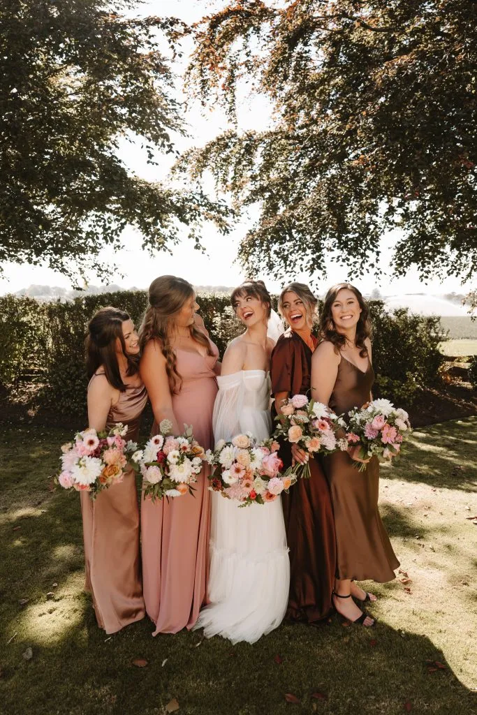 fraoch floral design bride in a white gown with her bridesmaids in blush and brown dresses, holding pastel bouquets outdoors under a tree.