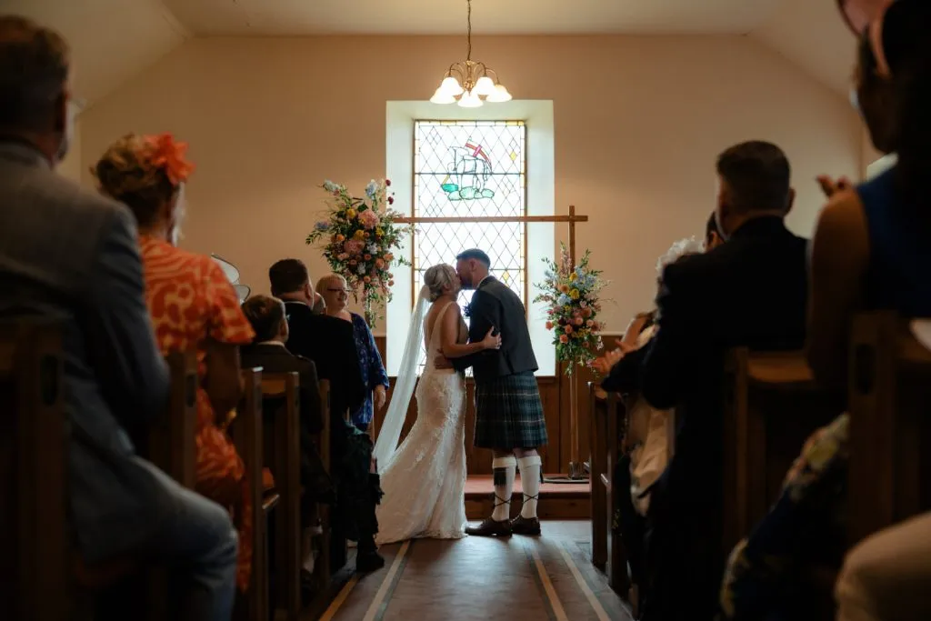 fraoch floral design bride and groom kiss at the altar during a church wedding, with a stained-glass window behind them and floral arches on either side. aisle seats and guests watch.