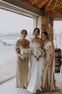 fraoch floral design bride in a strapless white gown stands between two bridesmaids in champagne dresses, all smiling and holding white bouquet arrangements of roses and baby's breath inside a rustic venue by a large window.