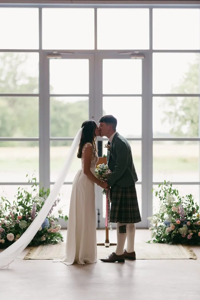 fraoch floral design bride and groom kiss indoors during their wedding, she holds a bouquet and her veil drapes behind them by tall windows with floral arrangements on the floor.