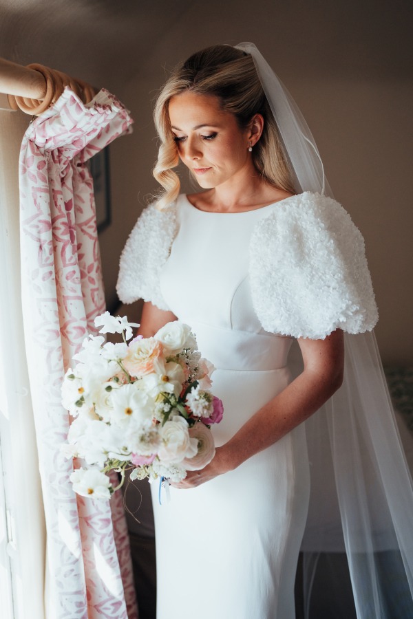 fraoch floral design bride in a white wedding gown with fluffy sleeves stands by a window, holding a pastel bouquet and looking down.