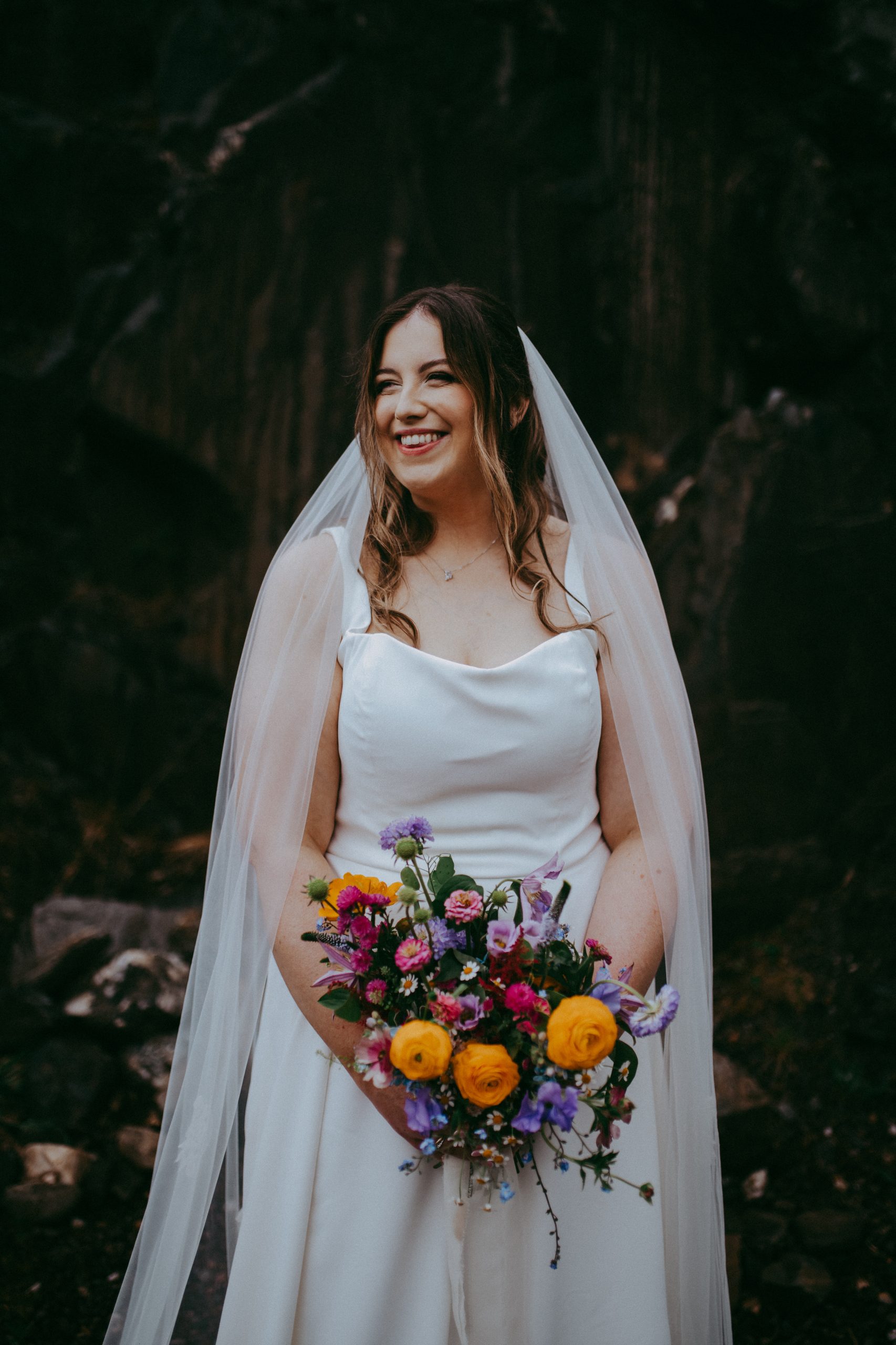 fraoch floral design bride in a white wedding dress and veil, smiling while holding a colorful bouquet outdoors.